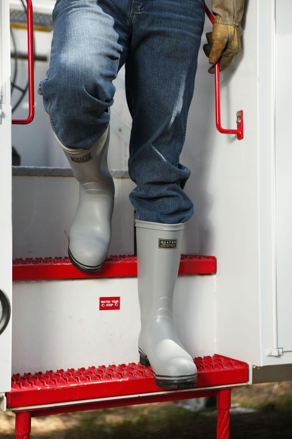 A person wearing FQD8 Series Quatro Dielectric Regular Boots descends red steps from a white vehicle. Only their lower body is visible, dressed in jeans and a glove on one hand. The setting appears to be outdoors.